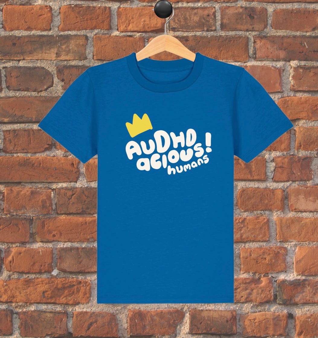 Blue t-shirt with white text and yellow crown graphic on a wooden hanger against a brick wall.