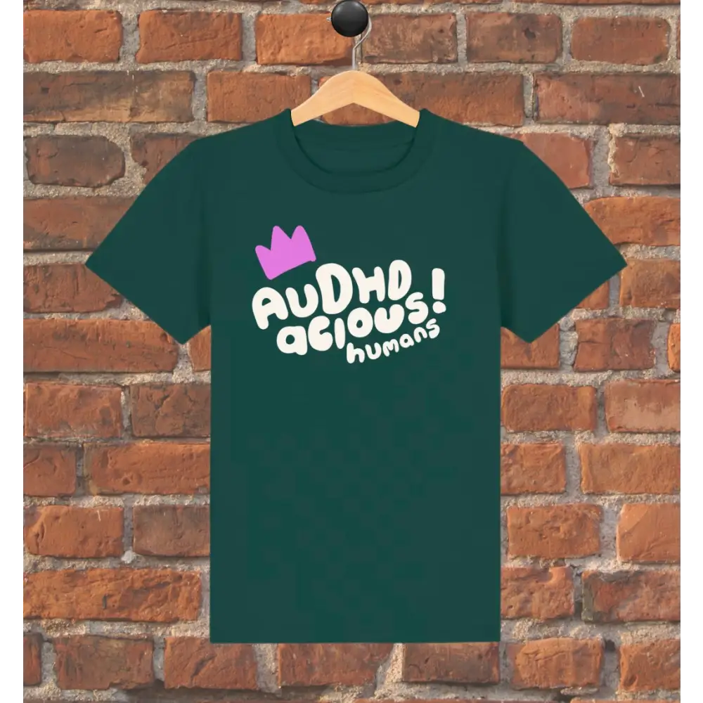 Green t-shirt with 'AUDHD delicious humans' text and purple crown on a wooden hanger against a brick wall.