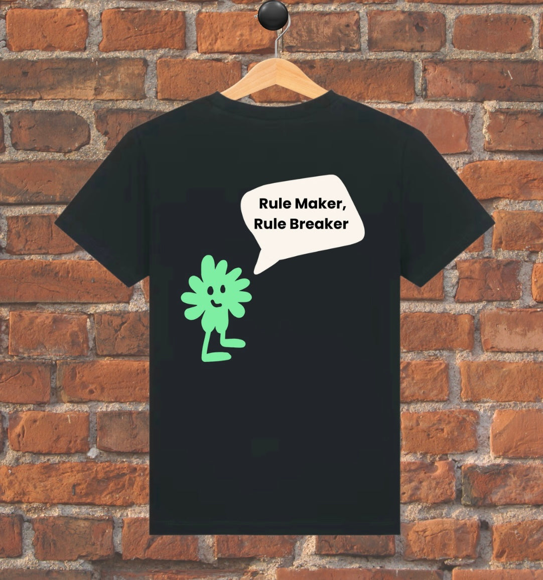 Black t-shirt with green character and text 'Rule Maker, Rule Breaker' on a wooden hanger against a brick wall.