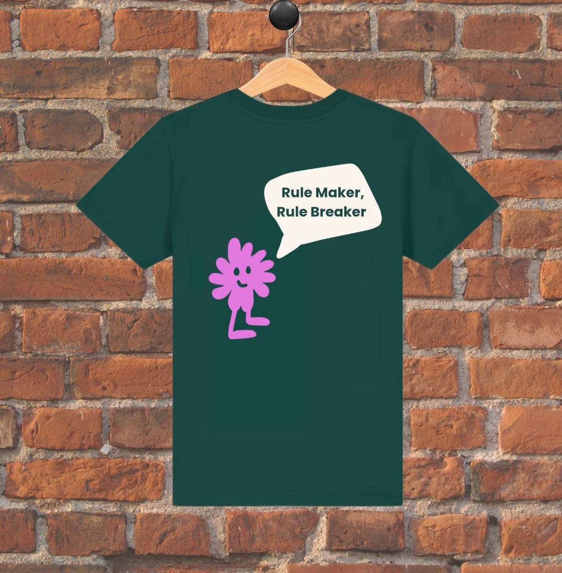 Green t-shirt with a purple flower and text on a wooden hanger against a brick wall.