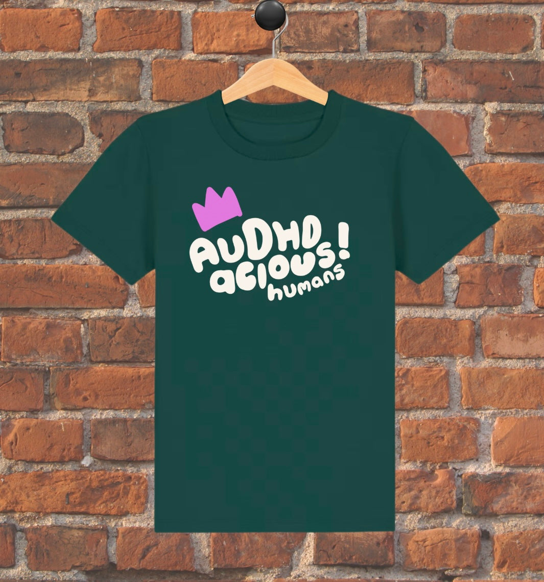 Green t-shirt with 'AUDHD delicious humans' text and purple crown on a wooden hanger against a brick wall.