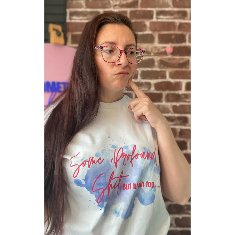 Person wearing a white t-shirt with colorful text and graphics, pointing to their head in front of a brick wall.
