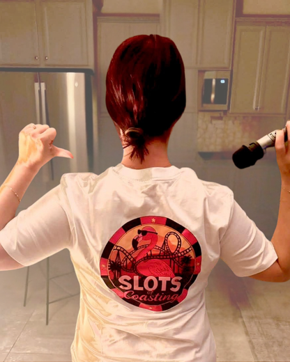 Amanda  from 'By Manna travel & Lifestyle' instagram shown from behind wearing a white t-shirt with a circular 'Slots of coasting' graphic on the back, standing in a kitchen and holding a microphone.
