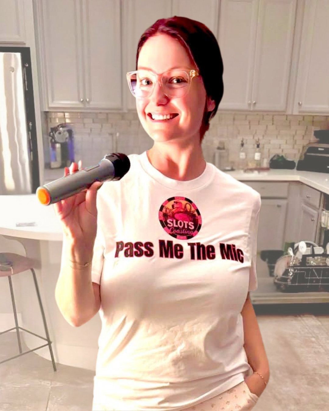 Amanda from 'By Manna travel & Lifestyle' instagram wearing clear glasses and a white graphic t-shirt stands in a kitchen holding a toy microphone. The t-shirt features a circular red and black graphic with the word 'SLOTS' and the text 'Pass Me The Mic' in dark red below it.