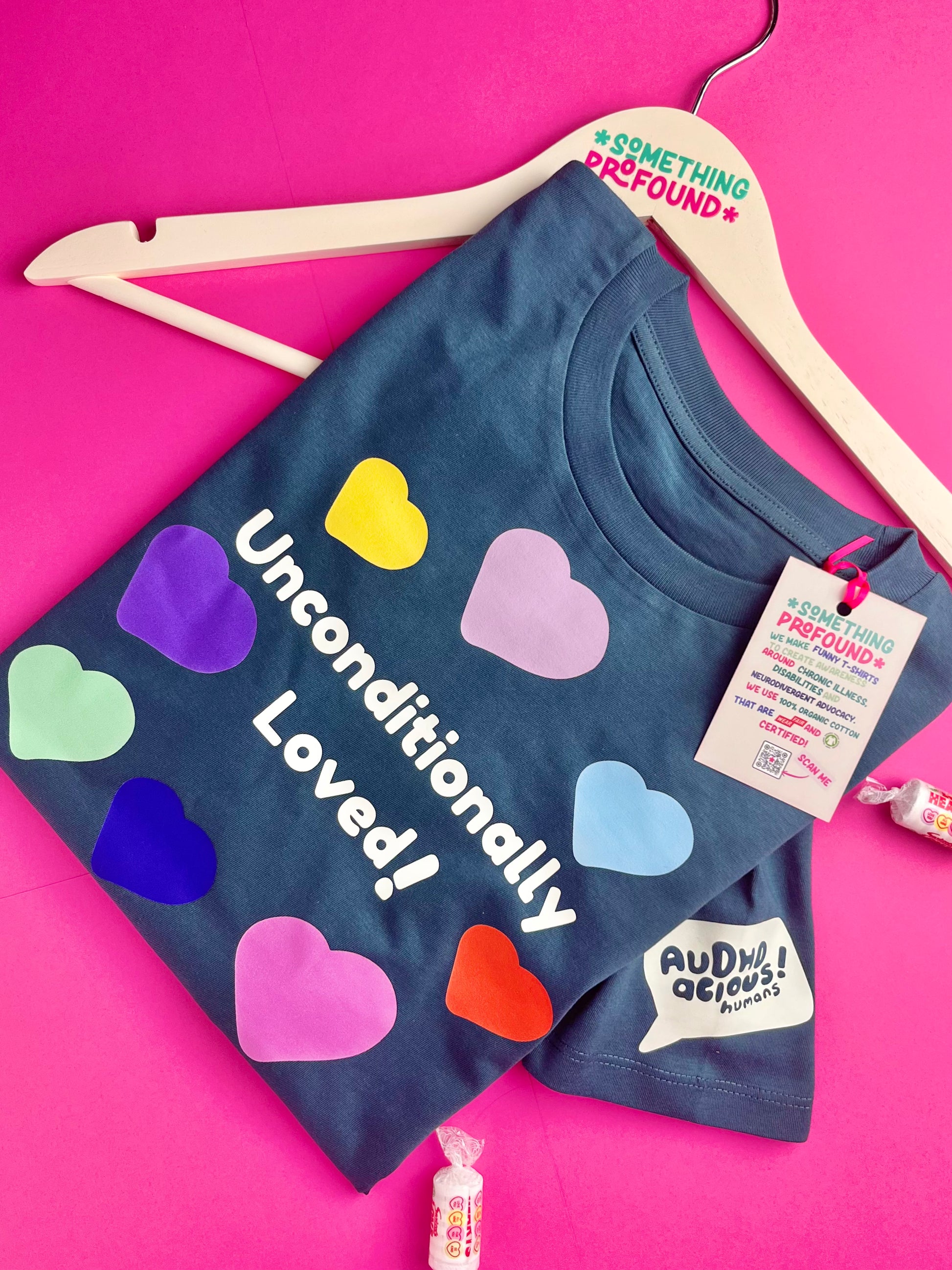 A stargazer t-shirt with 'Unconditionally Loved!' printed in white, decorated with colorful heart stickers, shown on a wooden hanger against a bright pink background.