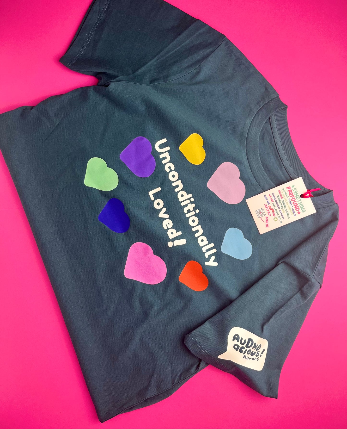 A dark teal t-shirt laid flat on a bright pink background, featuring the words 'Unconditionally Loved!' in white next to a cluster of colorful heart shapes in green, purple, yellow, pink, blue, and orange. A white tag and a sleeve patch both show the brand name 'Audie Devos'.