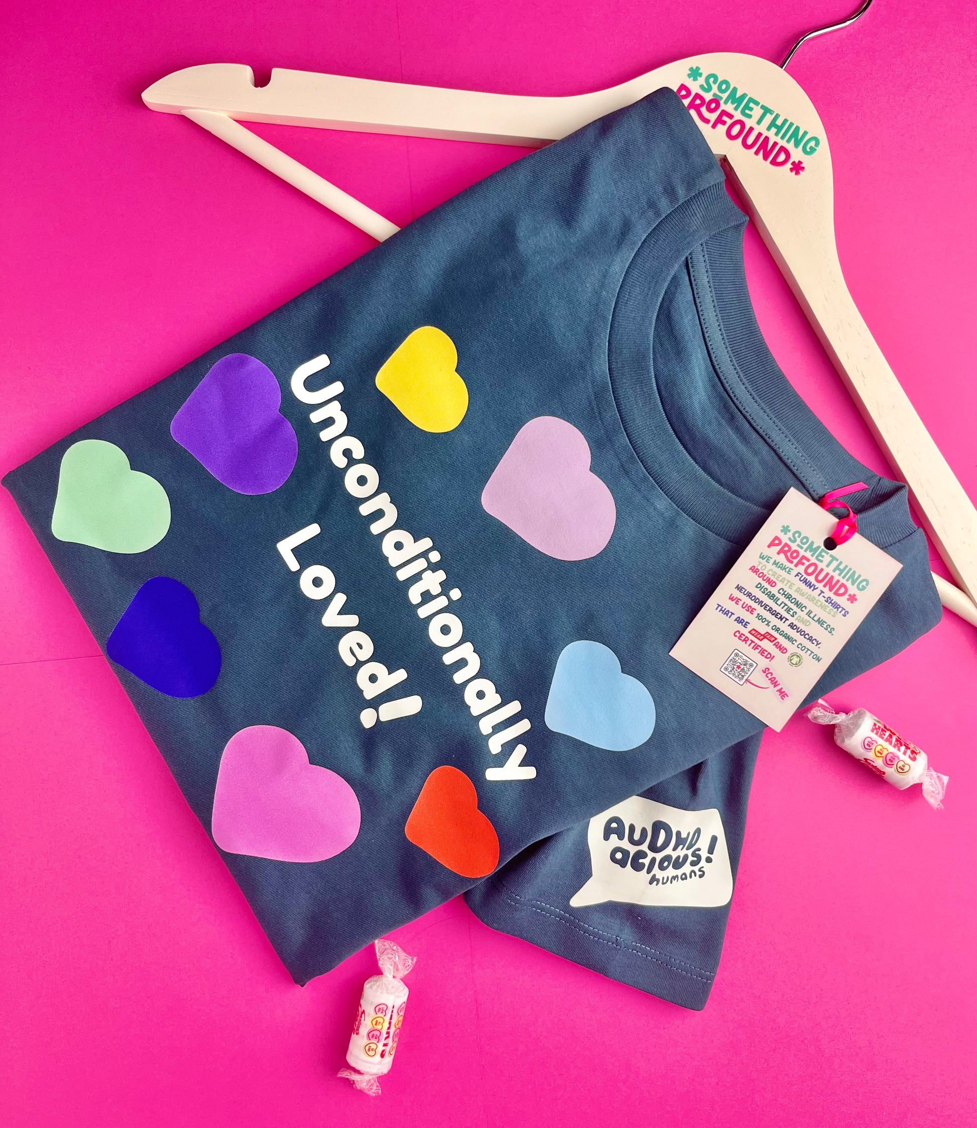 A teal graphic t-shirt with colorful heart shapes and the text 'Unconditionally Loved!' displayed on a light wood hanger against a bright pink background. The brand 'Ardment! Aesthetic Apparel' is visible on the hangtag and logo patch.
