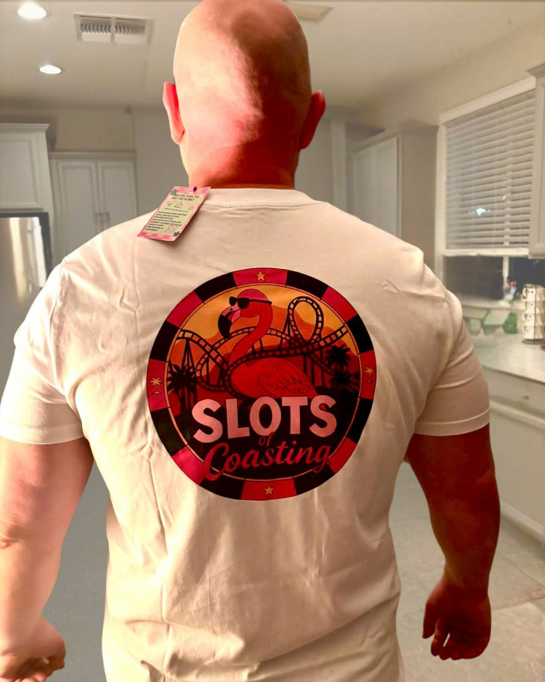 Dave from 'By Manna travel & Lifestyle' instagram viewed from behind wearing a white t-shirt with a large circular graphic on the back. The graphic features a red flamingo, a roller coaster, and the text 'SLOTS of Coasting' in white and pink, set against a kitchen background.