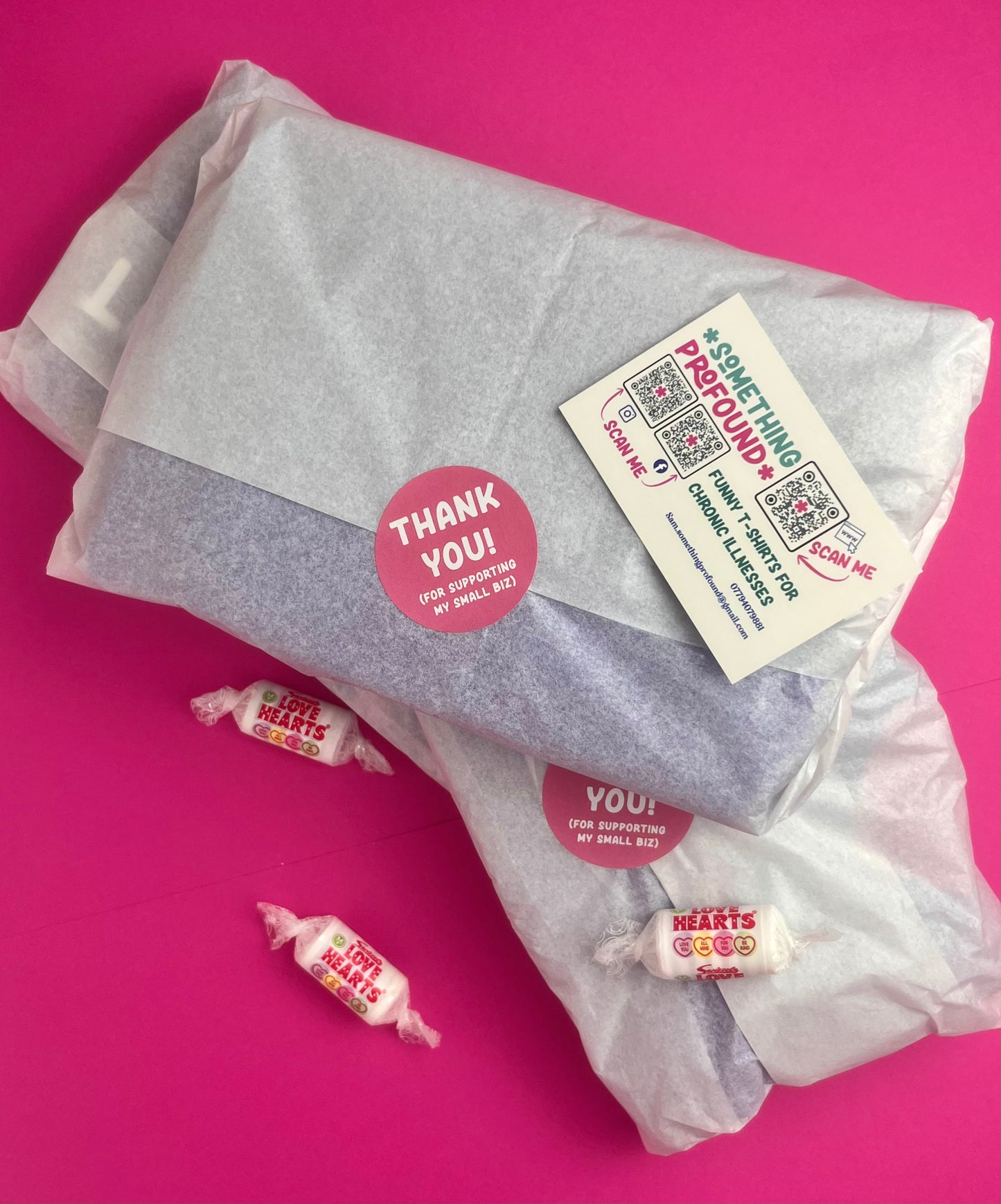 A gift-wrapped package wrapped in translucent tissue paper with a round pink 'Thank You!' sticker and a white something profound business card. three small wrapped love heart sweets are placed on a bright pink background.