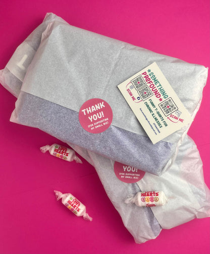 A gift-wrapped package wrapped in translucent tissue paper with a round pink 'Thank You!' sticker and a white something profound business card. three small wrapped love heart sweets are placed on a bright pink background.