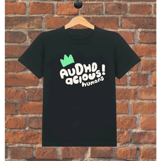 Black t-shirt with 'AUDHDacious humans!' text and green crown on a wooden hanger against a brick wall.