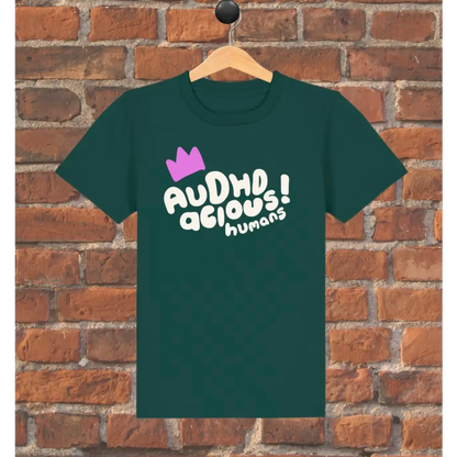 Green t-shirt with 'AUDHD delicious humans' text and purple crown on a wooden hanger against a brick wall.