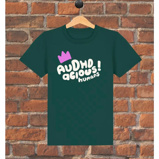 Green t-shirt with 'AUDHD delicious humans' text and purple crown on a wooden hanger against a brick wall.