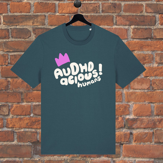 Green t-shirt with 'AUDHD delious humans' text and pink crown on a hanger against a brick wall.