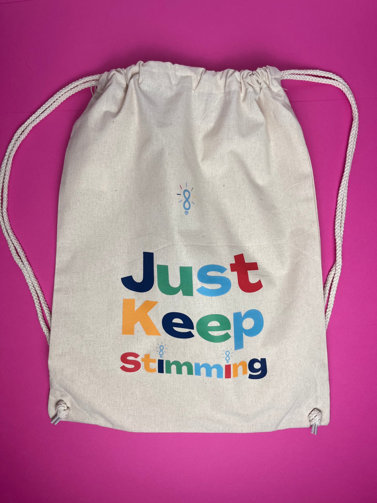 An off-white canvas drawstring bag with white rope drawstrings, featuring the colorful text 'Just Keep Stimming' printed on the front, set against a solid bright pink background.