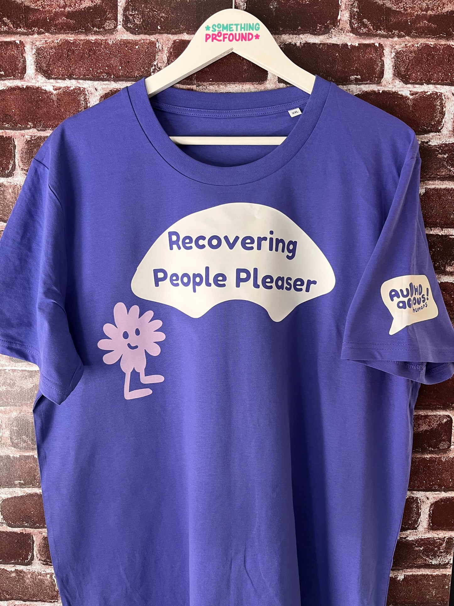 Blue t-shirt with 'Recovering People Pleaser' text on a hanger against a brick wall.