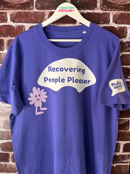 Blue t-shirt with 'Recovering People Pleaser' text on a hanger against a brick wall.