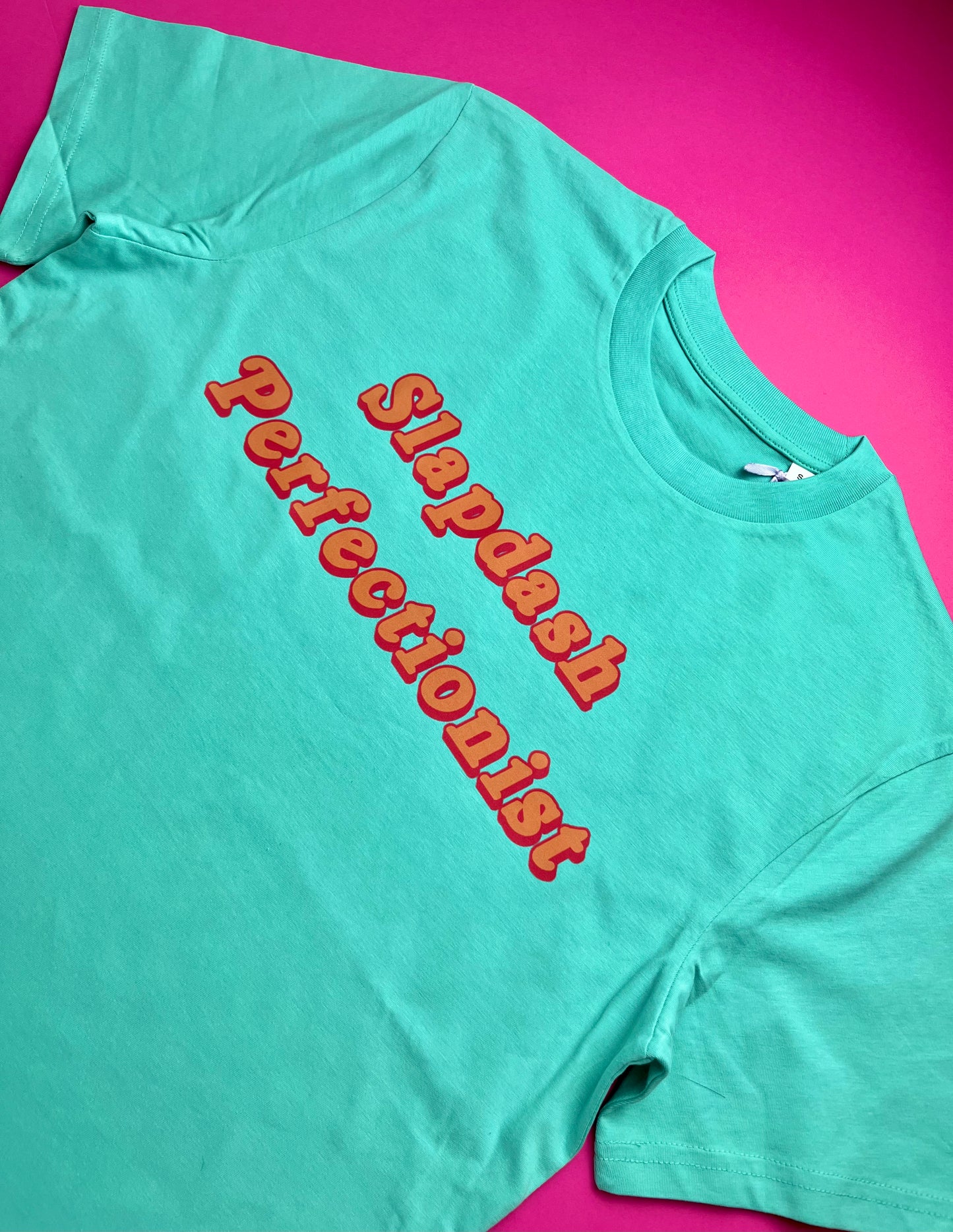 A pool blue t-shirt with 'Slapdash Perfectionist' printed in red 3D-effect letters, laid flat on a bright pink background.