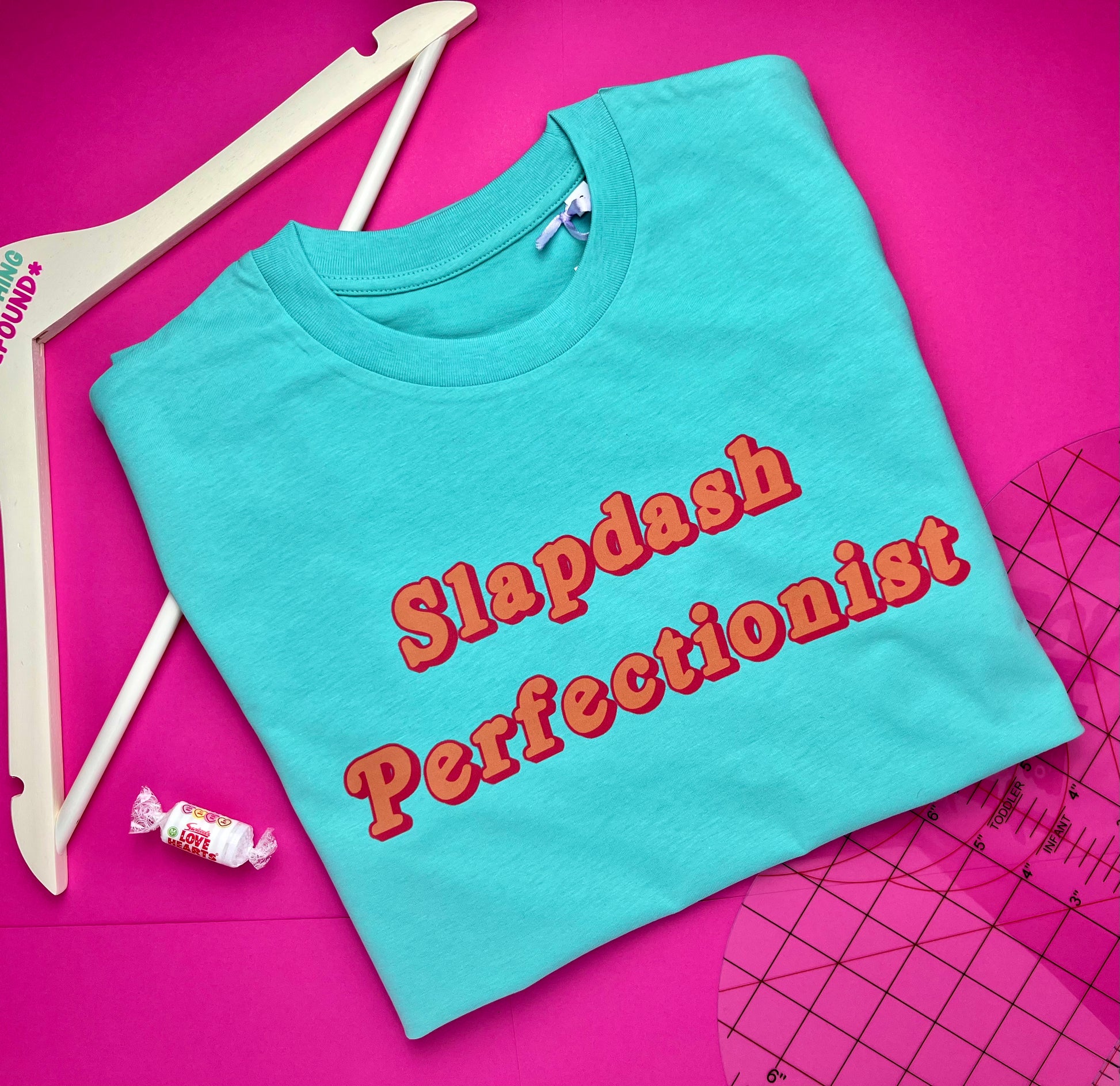 A pool blue folded T-shirt with the words 'Slapdash Perfectionist' in red outlined lettering, styled on a bright pink background with a coat hanger, ruler and a small pack of love hearts nearby.