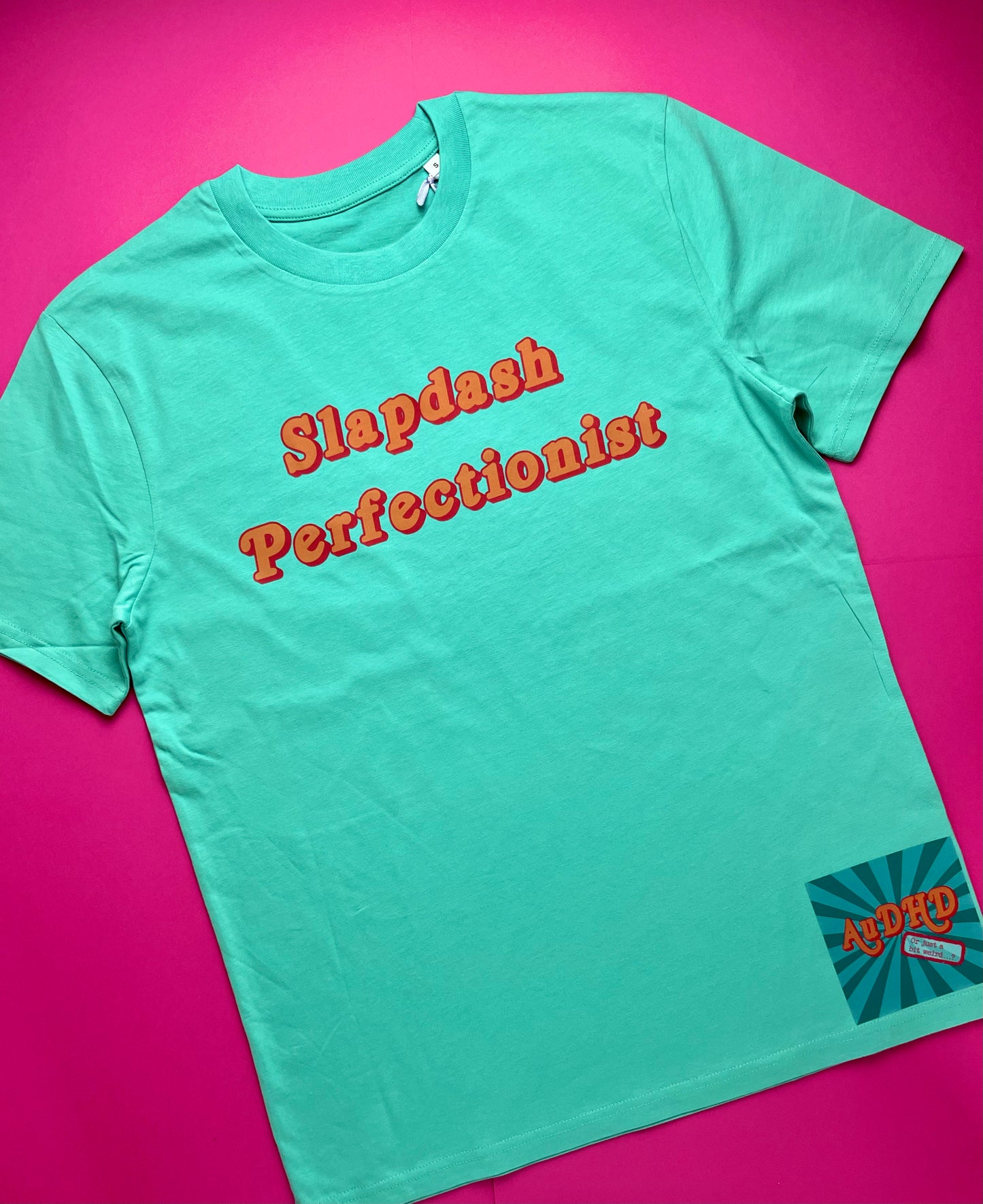 A pool blue short-sleeve t-shirt with 'Slapdash Perfectionist' printed in orange across the chest, laid flat on a vibrant pink background. A small retro-style 'AuDHD or just a bit weird' brand patch is visible on the bottom right hem.