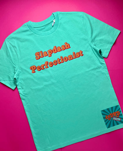 A pool blue short-sleeve t-shirt with 'Slapdash Perfectionist' printed in orange across the chest, laid flat on a vibrant pink background. A small retro-style 'AuDHD or just a bit weird' brand patch is visible on the bottom right hem.
