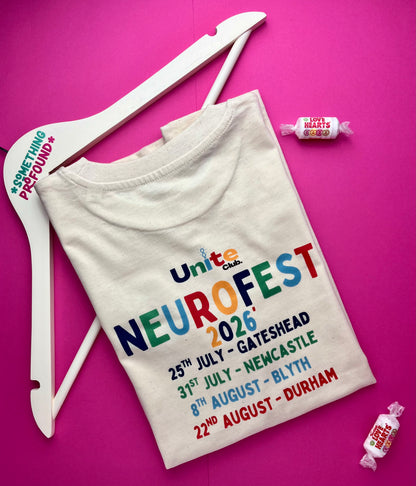 A folded cream-colored t-shirt with a colorful 'Unite Club NeuroFest 2026' event print, displayed on a white wooden hanger against a vibrant pink background with small candy props.