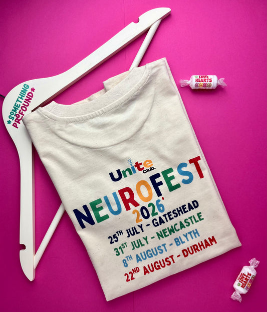 A folded cream-colored t-shirt with a colorful 'Unite Club NeuroFest 2026' event print, displayed on a white wooden hanger against a vibrant pink background with small candy props.