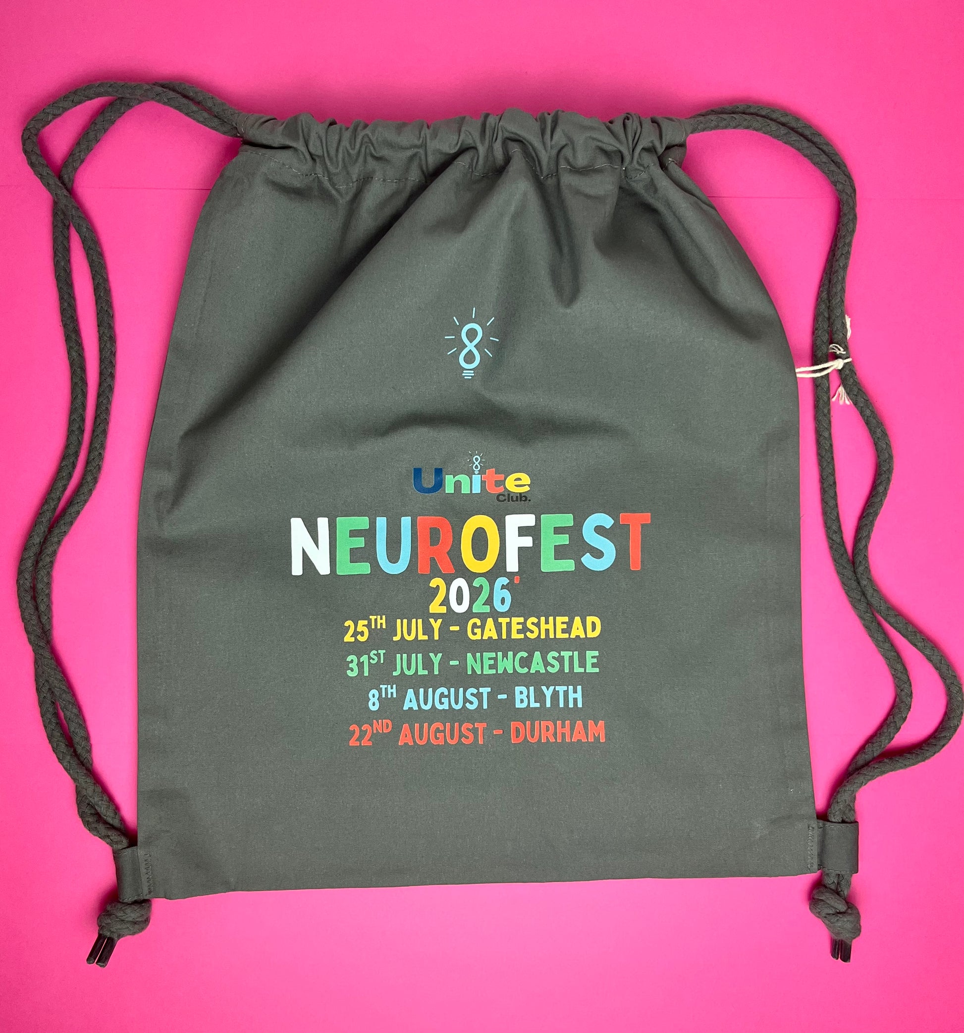 A dark gray drawstring backpack with 'Unite NeuroFest 2026' printed on the front, laid flat on a bright pink background.