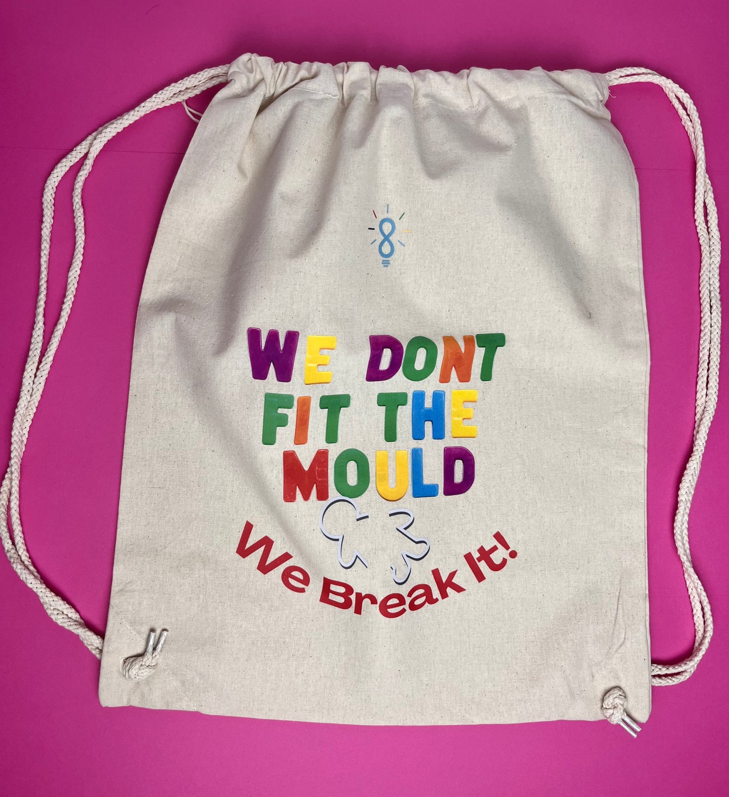 An off-white canvas drawstring bag with colorful text reading 'WE DONT FIT THE MOULD We Break It!' printed on the front, set against a solid magenta background.