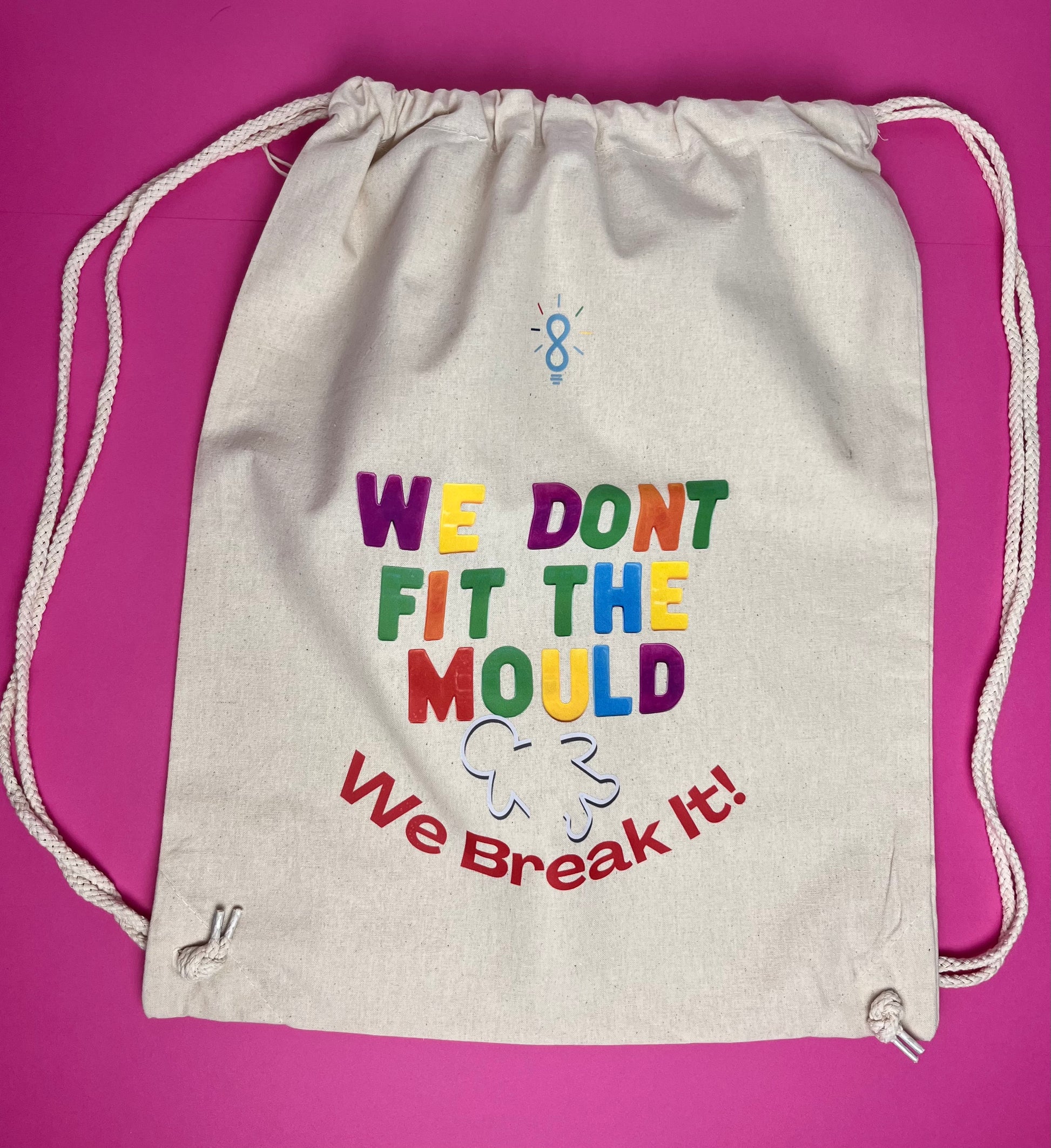 An off-white canvas drawstring bag with colorful text reading 'WE DONT FIT THE MOULD We Break It!' printed on the front, set against a solid magenta background.