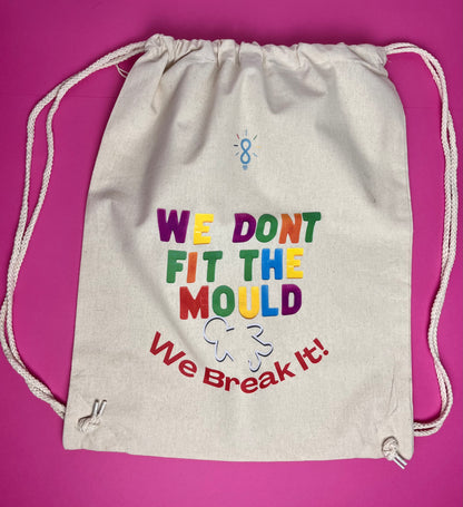 An off-white canvas drawstring bag with colorful text reading 'WE DONT FIT THE MOULD We Break It!' printed on the front, set against a solid magenta background.