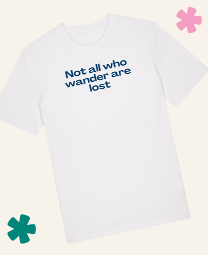 A white short-sleeve crewneck t-shirt with the phrase 'Not all who wander are lost' printed in blue text on the chest, displayed on a light cream background with decorative pink and teal flower graphics. Unite Club x Something Profound